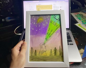 A hand holding up a framed child's crayon and colored-pencil drawing depicting a night sky, a moon, and trees, with a green sticky note on the glass reading 'Bon Festival Eve'.