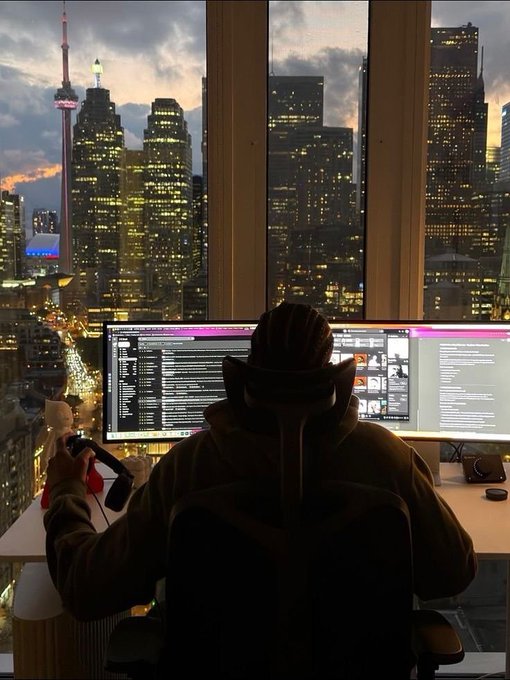 A person wearing a hoodie sits at a multi-monitor setup, with the CN Tower and a city skyline visible through the large window behind them.