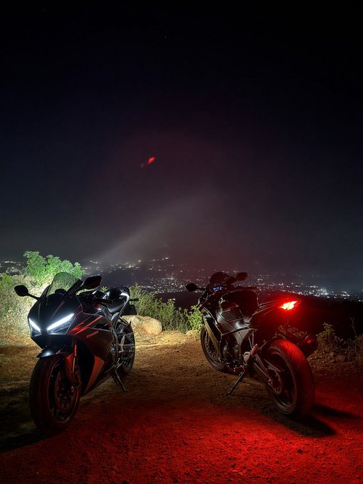 Two motorcycles parked on a dirt overlook at night, illuminated by their headlights and red tail light, with a sprawling city lights view below.