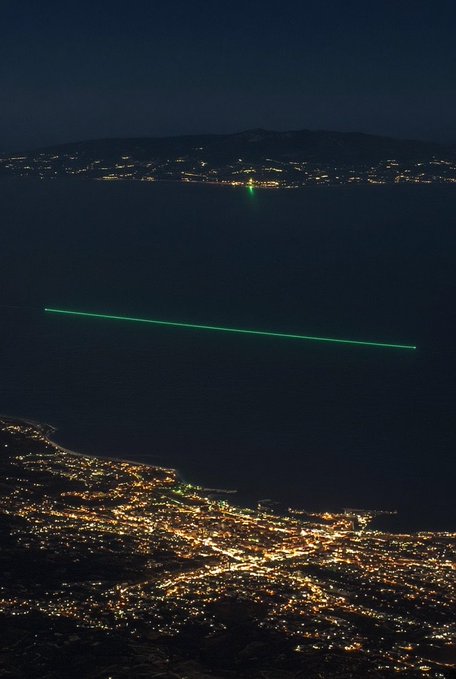 Nighttime aerial photograph of a coastal city with sparkling lights below and a bright green laser beam cutting horizontally across the dark sea toward a distant lighthouse.
