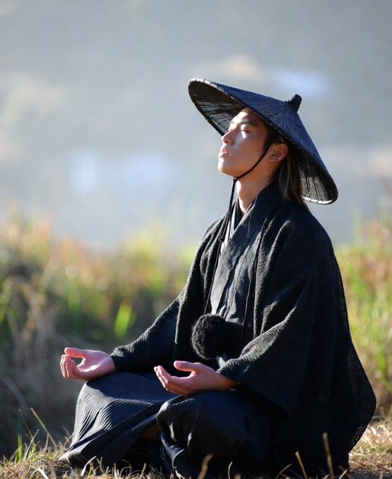 The image features an Asian man wearing traditional clothing such as a kimono and a hat, sitting cross-legged with his hands folded in front of him. The man appears to be meditating or taking a break from work. In the background, there is a body of water that adds to the serene atmosphere of the scene. The image also contains text that reads "you read the comments on ronin threads and it' s a dichotomy: scorned hearts and wallets, people who lost a lot of money investing in ronin and another group, those who were a bit later or not as invested early. they haven't been holding for years and watching their net worth". This text seems to be related to the man's attire and his location by the water, possibly suggesting that he is taking a break from work or meditation to enjoy the peaceful surroundings.