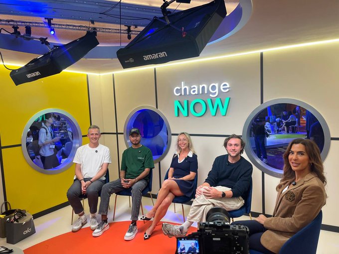 Five panelists sit on chairs in a brightly lit studio with yellow walls and circular windows, below a glowing 'change NOW' sign, with 'amaran' studio lighting overhead.