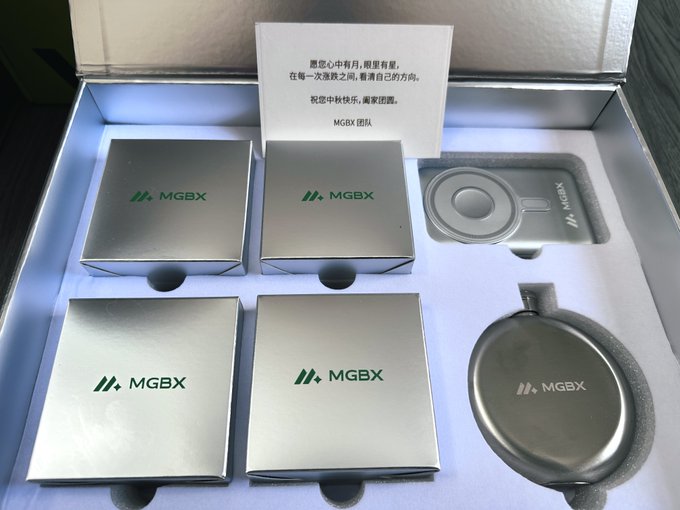An open MGBX gift box contains four silver boxes with the MGBX logo, a silver coin-shaped case, and a card with a Chinese message wishing the recipient a happy Mid-Autumn Festival.