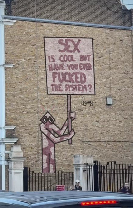 Street art painted on a brick wall depicts a figure holding a sign that reads 'SEX IS COOL BUT HAVE YOU EVER FUCKED THE SYSTEM?' with the signature '18ON8'.