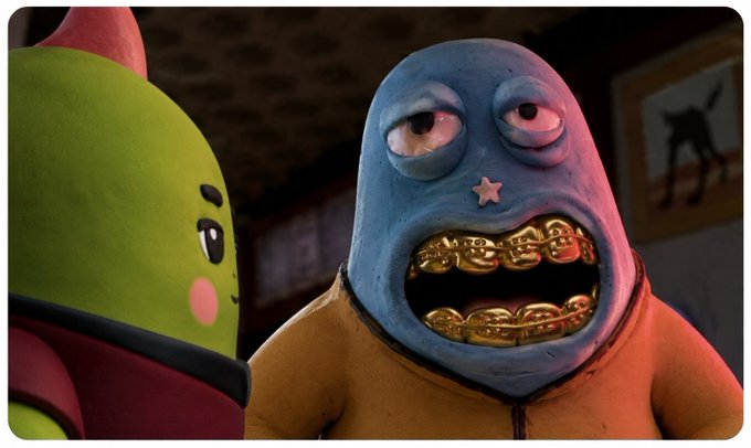 A close-up scene from a stop-motion animation showing a blue-skinned character with gold grills on his teeth and a star on his forehead looking at a green character.