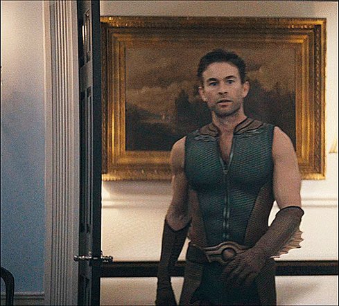 The image features a man dressed as a character from a comic book or movie, standing in front of a door with his arms crossed. He has a serious expression on his face, giving off an air of mystery. The background includes a painting hanging on the wall behind him and a chair positioned to the left side of the frame. There is also a post that says "Bitcoin and Ethereum when Dogecoin walks in". This suggests that there might be some connection between Bitcoin or Ethereum and the character he is portraying, possibly indicating that his costume represents a specific cryptocurrency or meme related to Bitcoin or Ethereum.