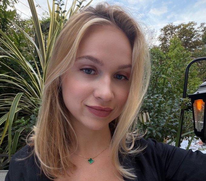 A blonde woman takes a selfie outdoors in a lush garden setting, wearing a delicate gold necklace with a green clover pendant and a black top, while a warm lantern glows softly behind her, complementing the post's theme of positive energy and personal connection.