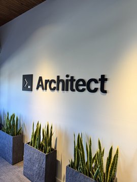The image shows the interior entrance signage for "Architect," featuring the company's logo and name displayed in bold black lettering on a light wall, complemented by potted snake plants in modern gray planters. This appears to be the new office or headquarters space referenced in the post as "the new home for trading AI compute," serving as a visual glimpse into Architect's physical location.