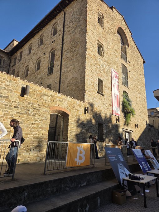 A historic stone building in Florence hosts a Bitcoin event, with a yellow "₿" banner and informational displays visible behind metal barriers, while a woman in a white mask walks past the setup. The juxtaposition of medieval architecture with cryptocurrency signage visually captures the theme of "digital Renaissance" blending tradition with modern fintech innovation.