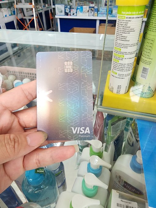 A hand holding a silver/pink metallic Visa Platinum credit card in front of store shelves containing various personal care products.