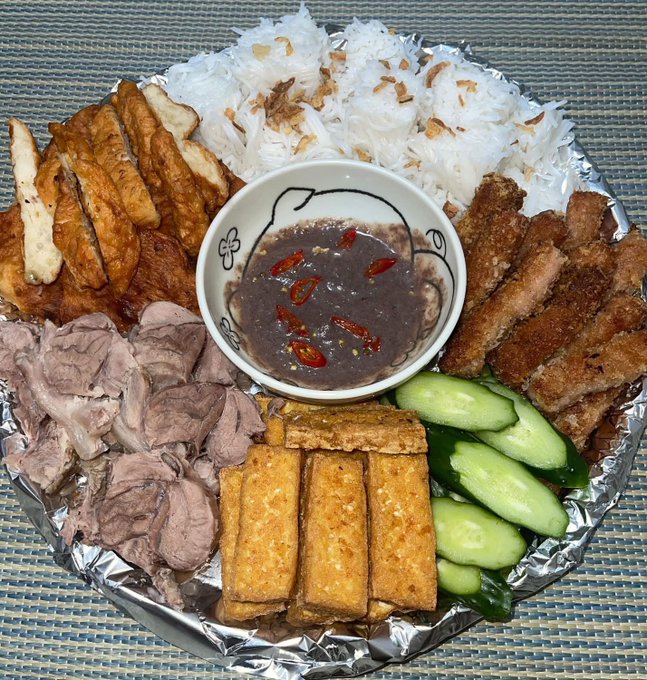 A platter of food lined with foil includes white rice, grilled chicken, fried pork, sliced beef, fried tofu or fish cakes, cucumber slices, and a central bowl of dark dipping sauce topped with red chili peppers.