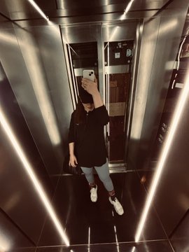 A person in a black shirt and jeans taking a mirror selfie with a smartphone in an elevator with metallic walls and lighting.
