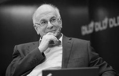 Black and white photograph of an elderly man with glasses, resting his chin on his hand in a thoughtful pose, with 'QUID' text visible in the blurred background.