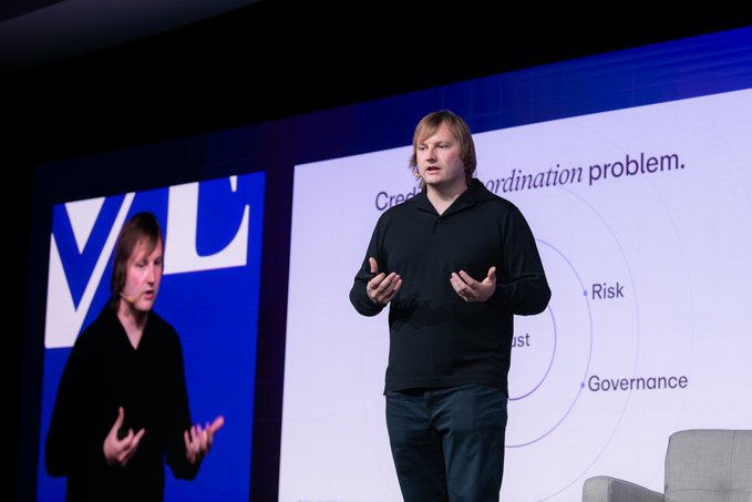 Two men on stage at a VC conference, with the speaker gesturing while discussing a coordination problem involving risk, trust, and governance shown on the presentation screen.