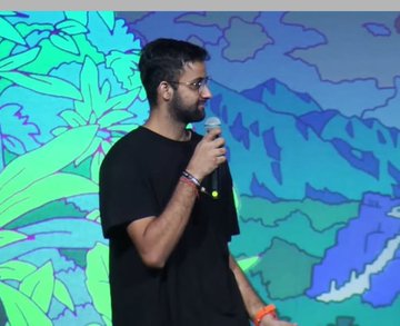 A man with a beard and glasses wearing a black t-shirt, speaking into a microphone while standing in front of a colorful illustrated backdrop with floral designs.