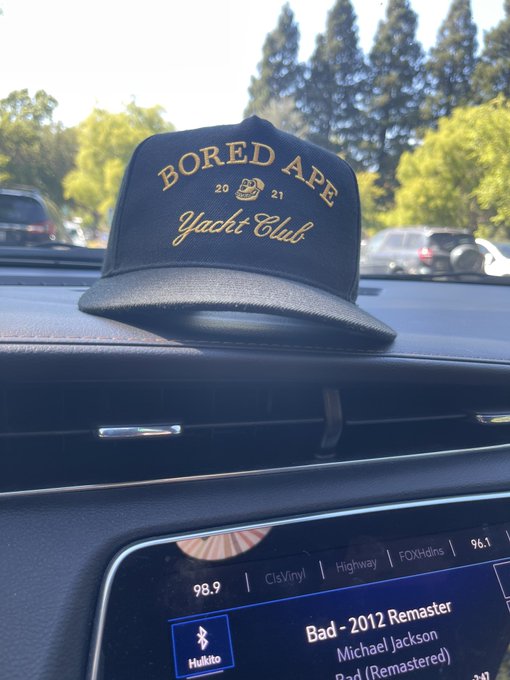 A black 'Bored Ape Yacht Club 2021' snapback hat on a car dashboard, with a radio display showing 'Bad - 2012 Remaster' by Michael Jackson playing.