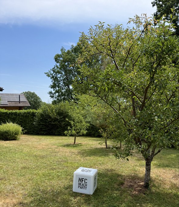A sunny outdoor scene with a small white cube labeled 'NFC BEACH PARTY' placed on grass near a young tree, with a house and solar panel visible in the background.
