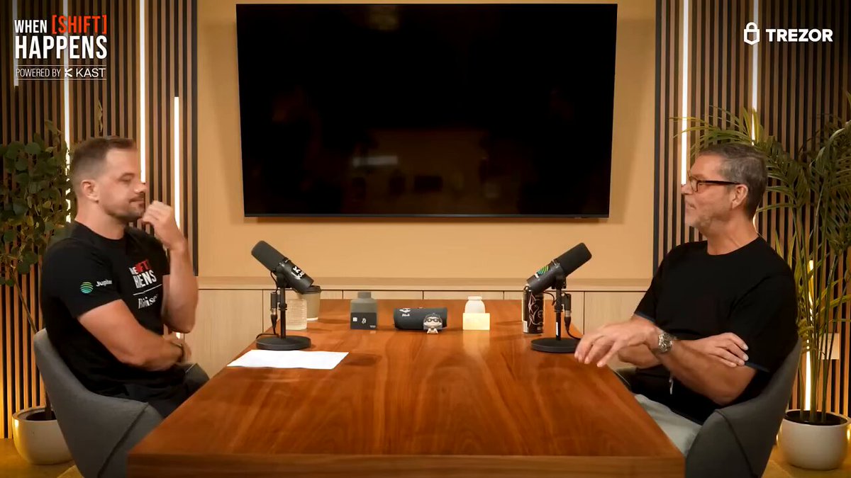 Two men are sitting at a wooden table, engaged in a conversation with microphones in front of them. The background features a television screen displaying a podcast episode or interview where they discuss Bitcoin as a trade opportunity. The man on the left has his arms crossed over his chest while the other man leans back in a chair, likely sharing their thoughts and opinions about this topic.