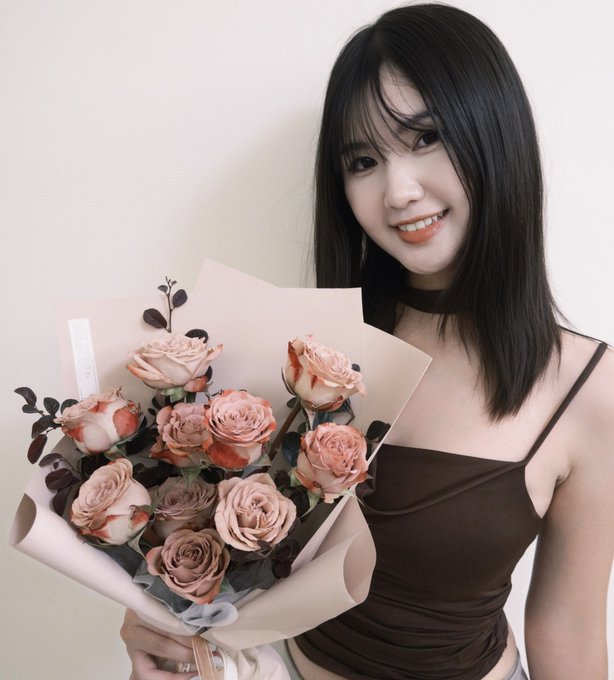 A young woman with long dark hair smiling while holding a bouquet of dusty pink roses wrapped in beige paper, wearing a brown top against a plain white background.