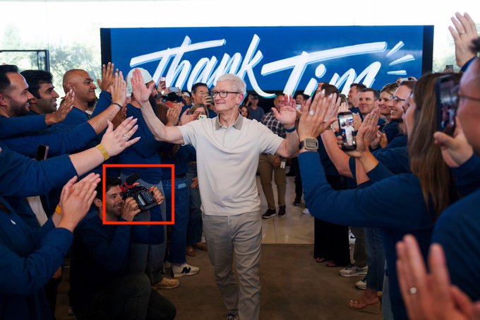 A man with glasses high-fives a crowd of cheering employees beneath a large blue "Thank Tim" banner, while a photographer highlighted in a red box captures the moment from the side—evoking the legendary, humble origin story of Tim Cook rising through Apple's ranks.