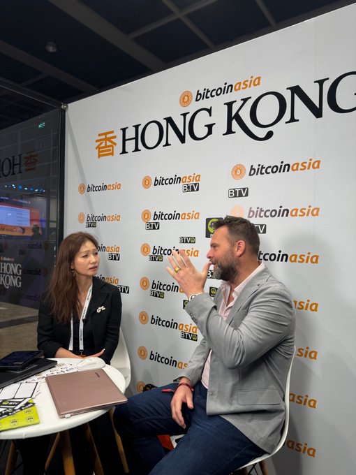 An interview at a Bitcoin Asia event in Hong Kong, with two people seated in front of a branded backdrop.