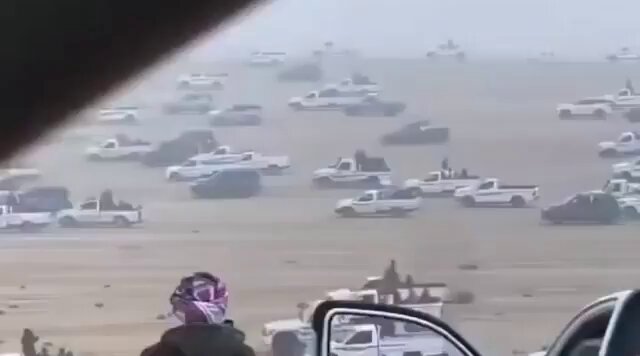 The image shows a large group of cars, including trucks and police vehicles, driving across a desert plain. There are at least 14 cars visible in various sizes and colors. The scene appears to be chaotic as people are scattered throughout the area, possibly fleeing or trying to escape from an event. One person is wearing a pink headscarf that stands out against the otherwise monotonous landscape of vehicles and sand.
