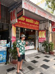 A man in a floral shirt and shorts stands on a sidewalk in front of a shop with red signage that reads 'DK Jeans' and 'SHOP Ngoài Lối'.
