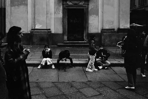 A black and white photograph showing several people on a city street in front of an old building, with one person on the left holding a camera and others sitting on the curb.