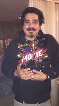 A man with a goatee wearing a black hoodie with 'MOXIE' text, holding an award trophy while standing in a kitchen.