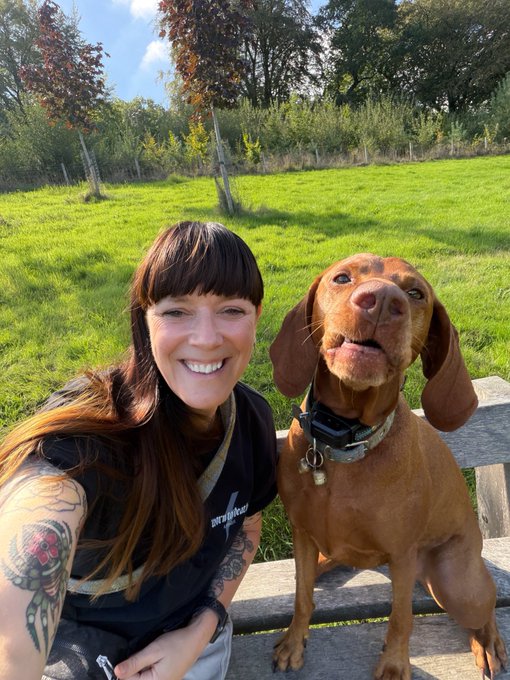 A smiling woman with dark hair and tattooed arms takes a selfie with a brown dog wearing a collar in a sunny grassy park setting.