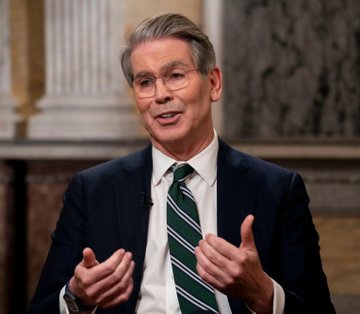A man with gray hair wearing glasses, a dark suit, and a green and white striped tie, gesturing with both hands while speaking in what appears to be a formal indoor venue.