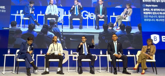 A panel discussion on stage at a conference with multiple seated speakers, including one identified by an on-screen overlay as 'Baylor Myers,' alongside live translation text in Korean and Japanese.