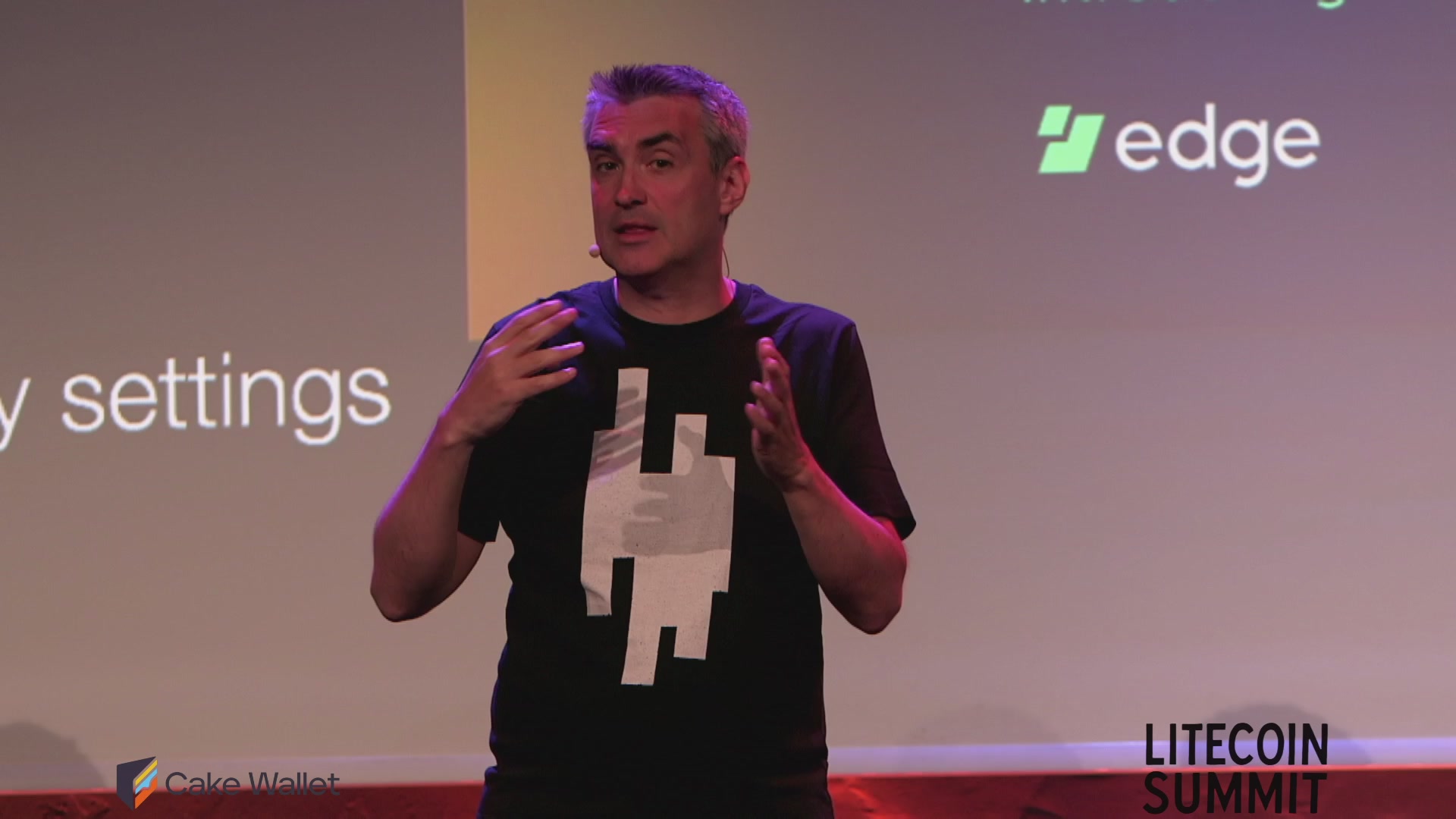 The image shows a man standing at a podium, giving a speech or presentation. He appears to be wearing a black t-shirt with white text that reads "Litecoin Summit". The background of the image features a large screen displaying the Litecoin Summit logo and other relevant information.