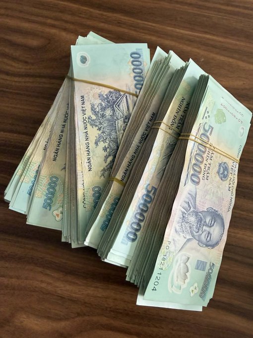Multiple stacks of Vietnamese đồng banknotes, including 50,000 and 500,000 denominations, bundled with rubber bands on a wooden surface.