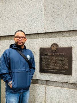 A man in a blue jacket stands next to a plaque at "55 Wall Street" in New York, humorously referencing the building as the source of late-night market volatility that triggers impulsive USDT buying and trading FOMO.