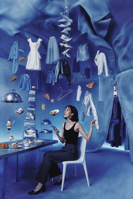 A woman in a black tank top and gray pants sits at a blue table enjoying breakfast while holding up a hanger with a silver jacket, surrounded by floating clothing, food, and accessories against a surreal blue backdrop.

The image plays on "gm" (good morning) by depicting an artistically styled morning scene where the woman is dressed and ready for the day, with food and outfits suspended in a dreamlike blue dreamscape that captures the whimsy of waking up.