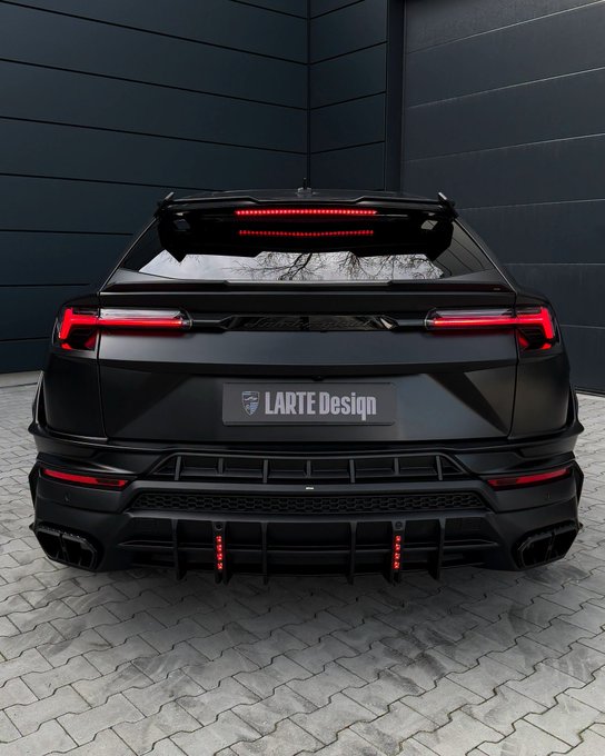 A rear view of a matte black Lamborghini Urus modified by LARTE Design, featuring a custom rear spoiler, aggressive diffuser with red accents, and quad exhaust tips, parked against a dark paneled wall.