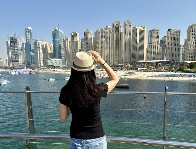A woman wearing a straw hat and black t-shirt gazes out at a sunlit marina lined with towering skyscrapers, a vibrant cityscape that mirrors the bullish, adventurous energy of "Cronos Summer" — embodying the post's confident, anonymous, on-the-go crypto vibe.