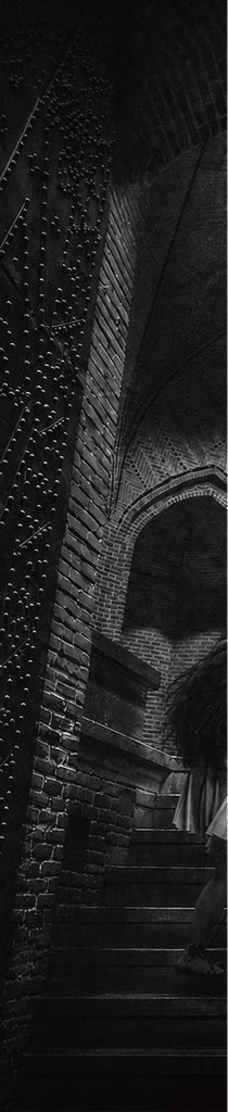 A moody black and white photograph of a figure in a white dress descending a dark staircase next to a textured brick archway.