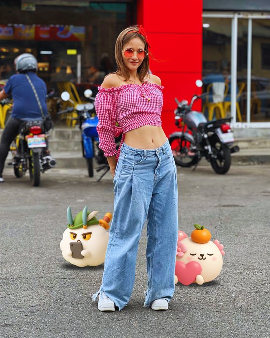 The image features a woman standing next to two cartoon characters, one of which has a face with an orange nose. The woman appears to be posing for a photo or participating in a contest related to the cartoon characters. The background shows a street scene with motorcycles parked nearby and other people present as well.