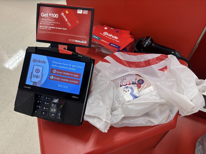 A Target checkout area featuring a payment terminal displaying a $100 gift card promotion, with a plastic shopping bag containing Disney's 'Wish' merchandise on the counter.