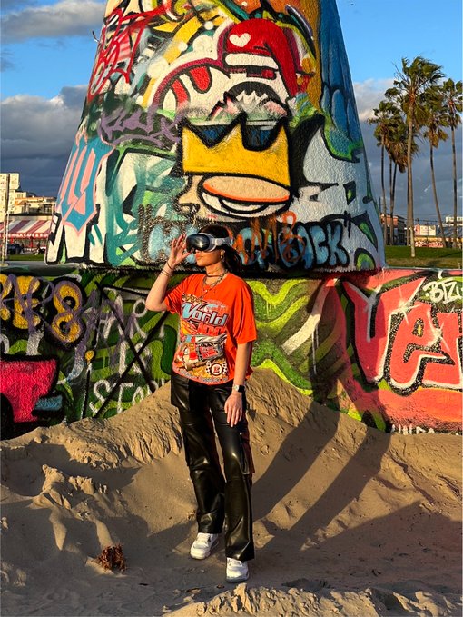 A person wearing VR goggles, an orange "World" t-shirt, and black pants stands on a sandy beach in front of a large graffiti-covered concrete structure.