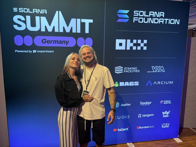 Two people posing in front of a 'Solana Summit Germany' backdrop featuring logos for Solana Foundation, OKX, Staking Facilities, VIA, Bags, Arcium, Jupiter, Seeker, and other crypto sponsors.