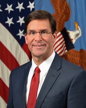 An official portrait of a man in a dark blue suit, white shirt, and red tie, wearing glasses, photographed in front of an American flag and a flag bearing a military eagle emblem.