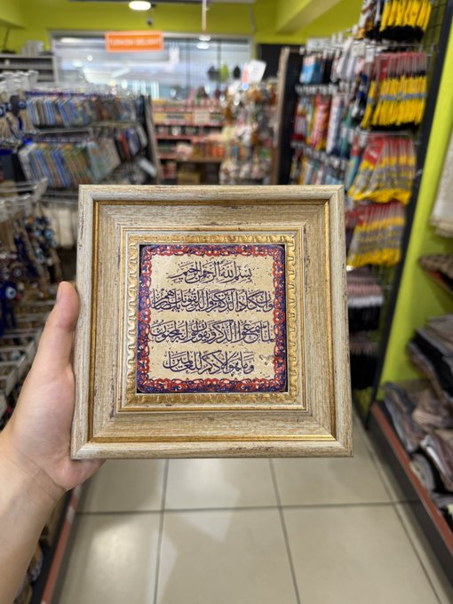 A hand holding a gold-framed piece of Islamic calligraphy art with Arabic script in a decorative tile design, photographed inside what appears to be a Middle Eastern gift or home goods shop.