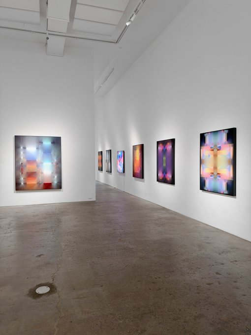 The image shows a spacious, minimalist gallery installation with white walls and polished concrete floors displaying a series of vibrant, abstract light-based artworks along both walls. The colorful pieces, featuring gradients of pink, purple, orange, and blue with prismatic, symmetrical patterns, appear to be the printed works from Zach Lieberman's "What Light May Be" solo exhibition, showcasing his exploration of light, color, and digital aesthetics.