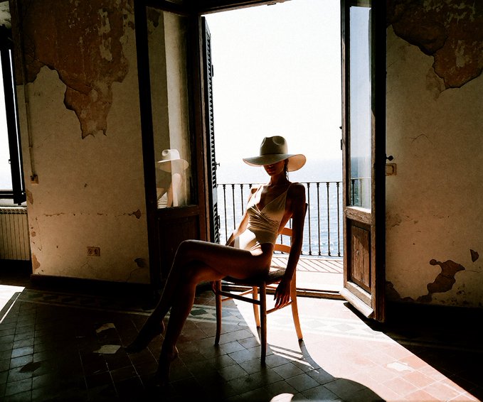 Artistic photograph of a woman wearing a white hat and tank top, seated on a wooden chair in a sunlit, weathered room with peeling walls, silhouetted against bright light streaming through an open doorway leading to a balcony with an ocean view.