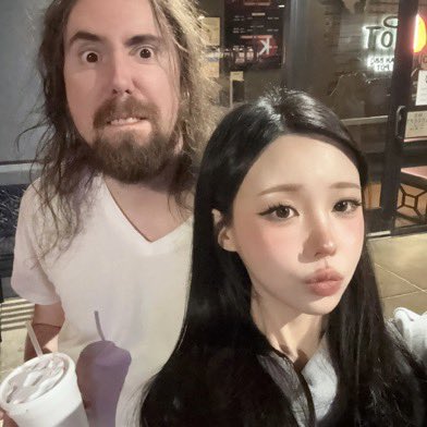 A selfie of a man with wild hair and a wide-eyed expression next to a young woman with long black hair and pouting lips, taken inside what appears to be a restaurant or cafe.
