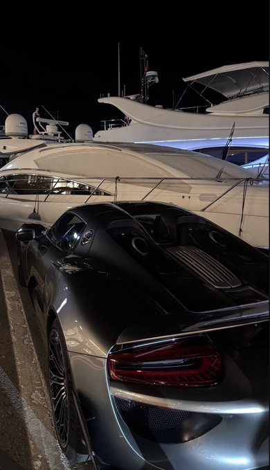 The image features a silver sports car parked next to a white yacht, both of which are docked at a marina or pier during nighttime hours. The car has its headlights on, illuminating the scene as it faces towards the right side of the photo. In the background, there is a boat visible in the distance, adding to the overall maritime atmosphere. The caption "Retire off $XRP" suggests that this image might be related to an advertisement or promotional material for Bitcoin or cryptocurrency exchanges.