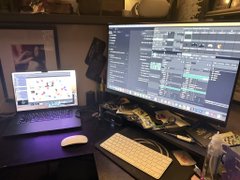 A desk setup featuring a dual-monitor workspace with a large external monitor displaying audio editing software, a laptop, an Apple Magic Mouse, a wireless keyboard, studio monitors on stands, and various audio equipment.
